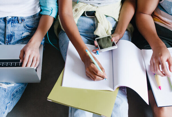 close-up-overhead-portrait-girl-blue-shirt-jeans-holding-laptop-knees-while-sitting-beside-university-mates-female-student-writing-lecture-notebook-using-phone-friends-e1649274050563